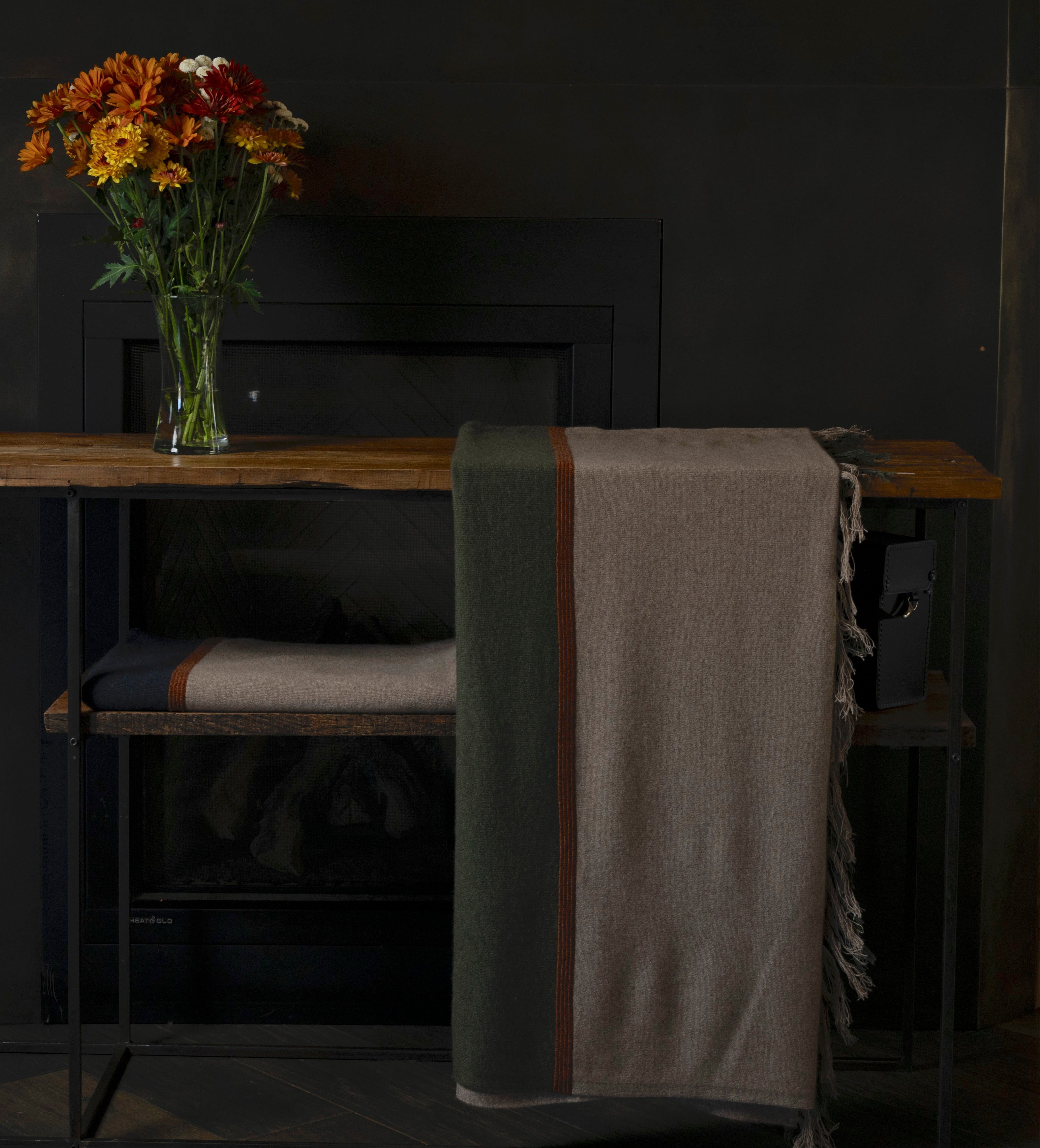 Wooden table with a vase of flowers and a blanket draped over a chair against a dark wall.